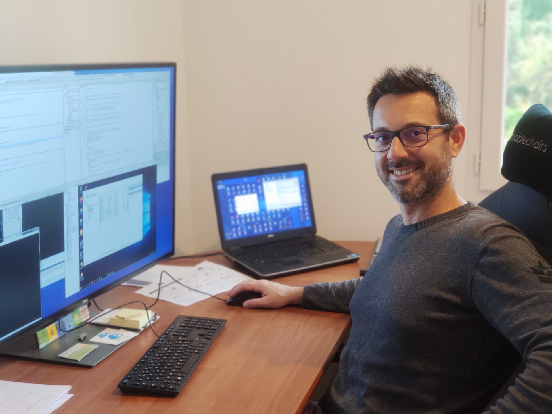 Un professionnel souriant travaillant sur plusieurs écrans dans un bureau, illustrant un environnement de travail informatique moderne.